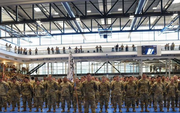 A large formation of military personnel in camouflage uniforms (OCP/MultiCam pattern) stands at attention inside a modern indoor sports facility or gymnasium. A central figure holds a unit guidon flag while rows of soldiers are arranged in precise formation on blue floor mats. Spectators and additional personnel observe from upper balcony levels surrounding the main floor.