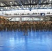 A large formation of military personnel in camouflage uniforms (OCP/MultiCam pattern) stands at attention inside a modern indoor sports facility or gymnasium. A central figure holds a unit guidon flag while rows of soldiers are arranged in precise formation on blue floor mats. Spectators and additional personnel observe from upper balcony levels surrounding the main floor.