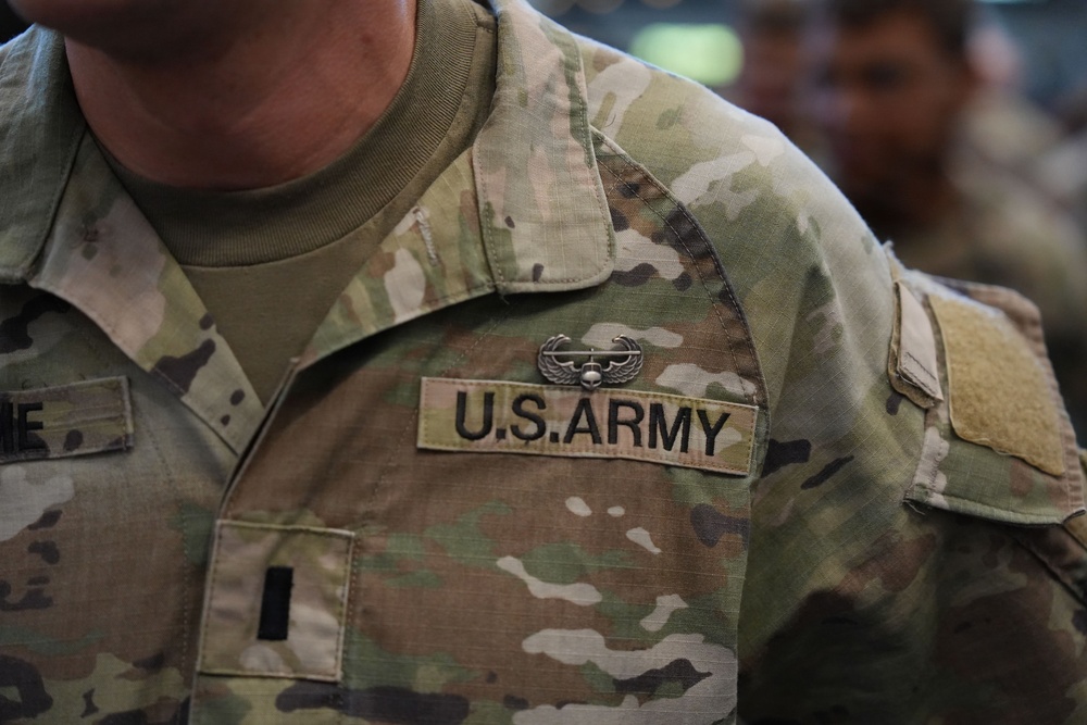 Close-up photograph of a U.S. Army soldier's upper torso wearing Operational Camouflage Pattern (OCP) uniform. The uniform displays a 'U.S. ARMY' chest tape, an Aviation Badge (Air Assault or similar wings insignia) above the tape, and a single black bar rank insignia indicating the rank of First Lieutenant or Second Lieutenant. Additional soldiers in similar uniforms are visible in the blurred background.