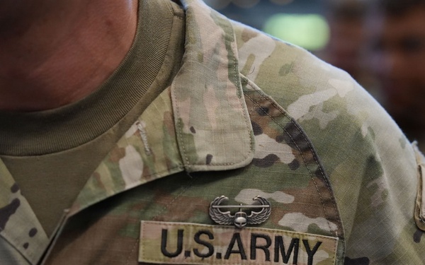 Close-up photograph of a U.S. Army soldier's upper torso wearing Operational Camouflage Pattern (OCP) uniform. The uniform displays a 'U.S. ARMY' chest tape, an Aviation Badge (Air Assault or similar wings insignia) above the tape, and a single black bar rank insignia indicating the rank of First Lieutenant or Second Lieutenant. Additional soldiers in similar uniforms are visible in the blurred background.