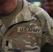 Close-up photograph of a U.S. Army soldier's upper torso wearing Operational Camouflage Pattern (OCP) uniform. The uniform displays a 'U.S. ARMY' chest tape, an Aviation Badge (Air Assault or similar wings insignia) above the tape, and a single black bar rank insignia indicating the rank of First Lieutenant or Second Lieutenant. Additional soldiers in similar uniforms are visible in the blurred background.