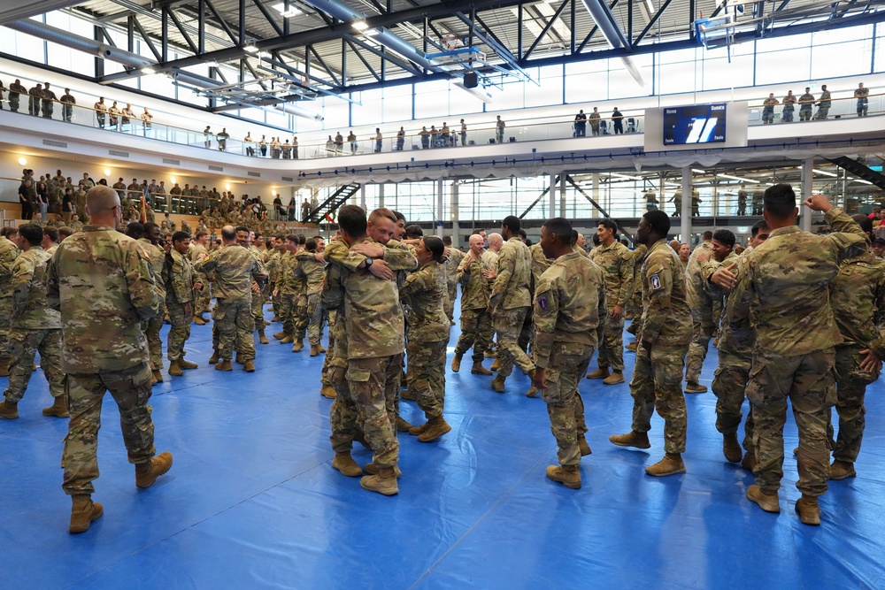 A large group of military personnel in US Army OCP (Operational Camouflage Pattern) uniforms are gathered inside a modern gymnasium or sports facility. Soldiers on the main floor are embracing and greeting one another, suggesting a reunion, homecoming, or post-ceremony gathering. Additional personnel are visible on upper-level balconies observing the event.