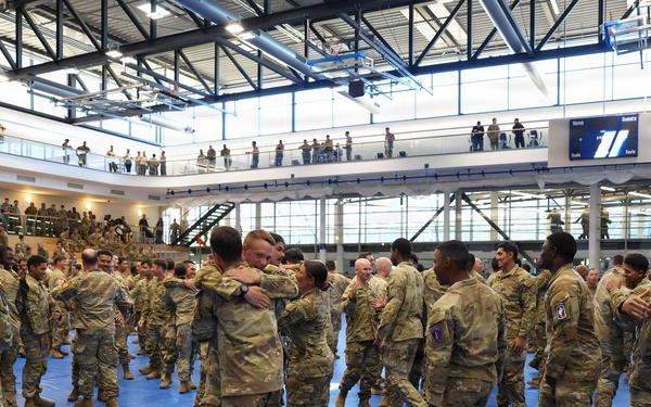 A large group of military personnel in US Army OCP (Operational Camouflage Pattern) uniforms are gathered inside a modern gymnasium or sports facility. Soldiers on the main floor are embracing and greeting one another, suggesting a reunion, homecoming, or post-ceremony gathering. Additional personnel are visible on upper-level balconies observing the event.