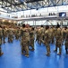 A large group of military personnel in US Army OCP (Operational Camouflage Pattern) uniforms are gathered inside a modern gymnasium or sports facility. Soldiers on the main floor are embracing and greeting one another, suggesting a reunion, homecoming, or post-ceremony gathering. Additional personnel are visible on upper-level balconies observing the event.