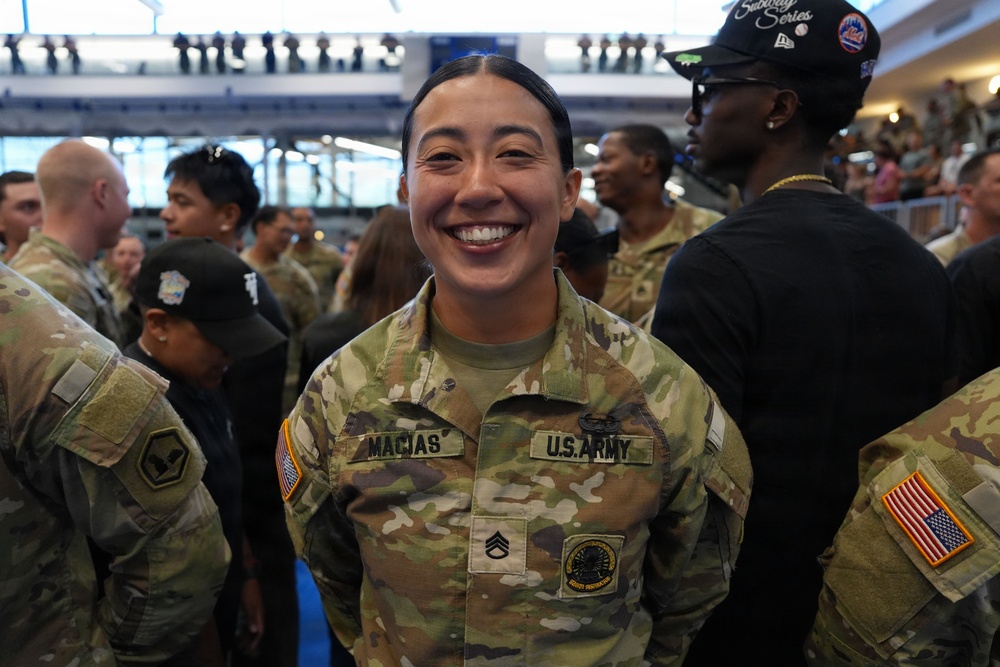 A U.S. Army soldier named Macias, wearing the rank of Staff Sergeant, smiles broadly at what appears to be a large indoor military event or ceremony. She is dressed in OCP (Operational Camouflage Pattern) uniform with U.S. Army tape, name tape, and various patches including an American flag on the sleeve. Multiple other soldiers and civilians are visible in the background of what appears to be a large venue or arena.