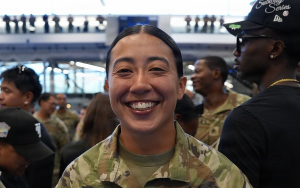 A U.S. Army soldier named Macias, wearing the rank of Staff Sergeant, smiles broadly at what appears to be a large indoor military event or ceremony. She is dressed in OCP (Operational Camouflage Pattern) uniform with U.S. Army tape, name tape, and various patches including an American flag on the sleeve. Multiple other soldiers and civilians are visible in the background of what appears to be a large venue or arena.