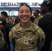 A U.S. Army soldier named Macias, wearing the rank of Staff Sergeant, smiles broadly at what appears to be a large indoor military event or ceremony. She is dressed in OCP (Operational Camouflage Pattern) uniform with U.S. Army tape, name tape, and various patches including an American flag on the sleeve. Multiple other soldiers and civilians are visible in the background of what appears to be a large venue or arena.