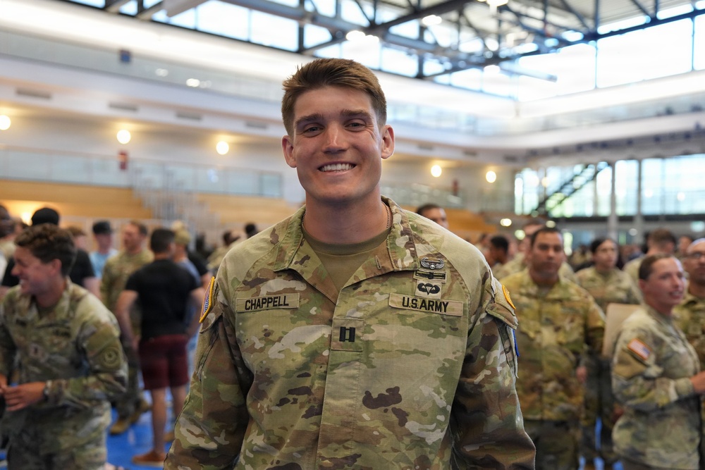 A U.S. Army Captain named Chappell is smiling at the camera while standing in what appears to be a large indoor facility. He is wearing the Operational Camouflage Pattern (OCP) uniform with visible rank insignia (Captain's bars) and a parachutist badge. Multiple other soldiers and civilians are visible in the background.