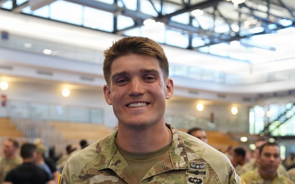 A U.S. Army Captain named Chappell is smiling at the camera while standing in what appears to be a large indoor facility. He is wearing the Operational Camouflage Pattern (OCP) uniform with visible rank insignia (Captain's bars) and a parachutist badge. Multiple other soldiers and civilians are visible in the background.