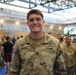 A U.S. Army Captain named Chappell is smiling at the camera while standing in what appears to be a large indoor facility. He is wearing the Operational Camouflage Pattern (OCP) uniform with visible rank insignia (Captain's bars) and a parachutist badge. Multiple other soldiers and civilians are visible in the background.