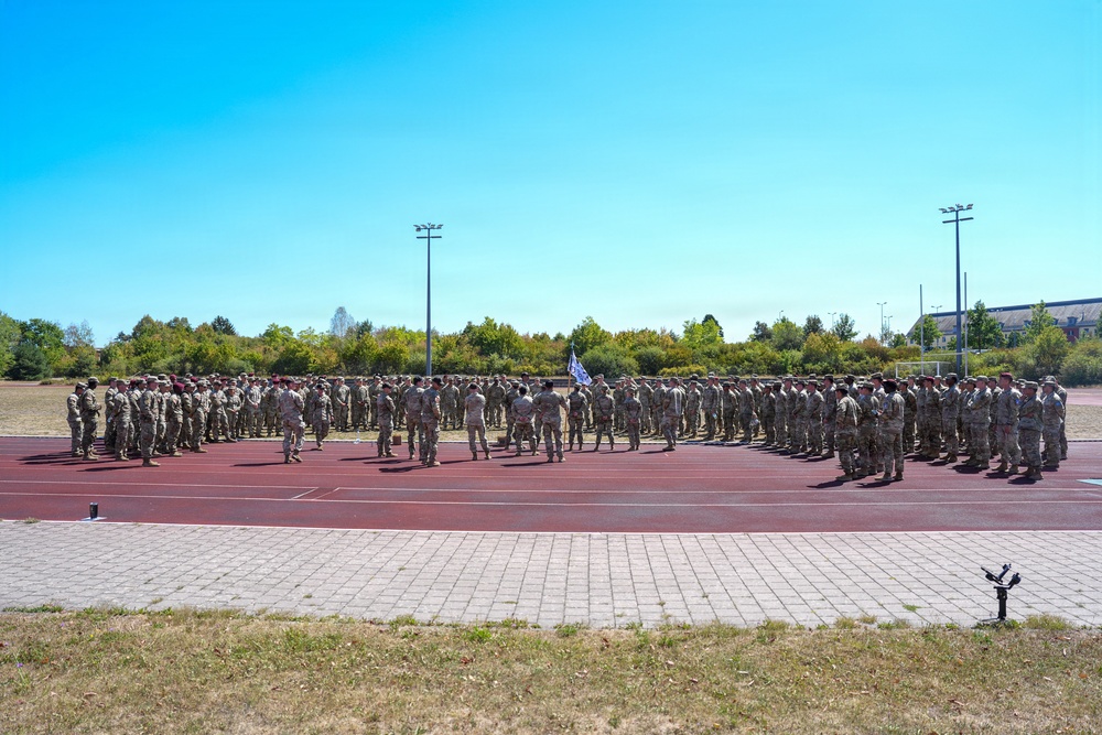 A large formation of military personnel in camouflage uniforms are assembled in a horseshoe or U-shaped formation on a red running track at what appears to be a military installation or sports facility. A flag or guidon is visible near the center of the formation, suggesting a formal military ceremony, briefing, or unit assembly. The event takes place outdoors under clear blue skies on a sunny day.