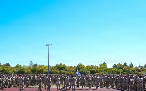 A large formation of military personnel in camouflage uniforms are assembled in a horseshoe or U-shaped formation on a red running track at what appears to be a military installation or sports facility. A flag or guidon is visible near the center of the formation, suggesting a formal military ceremony, briefing, or unit assembly. The event takes place outdoors under clear blue skies on a sunny day.