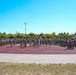 A large formation of military personnel in camouflage uniforms are assembled in a horseshoe or U-shaped formation on a red running track at what appears to be a military installation or sports facility. A flag or guidon is visible near the center of the formation, suggesting a formal military ceremony, briefing, or unit assembly. The event takes place outdoors under clear blue skies on a sunny day.