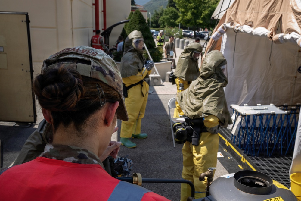 The image depicts what appears to be a HAZMAT or CBRN (Chemical, Biological, Radiological, Nuclear) training exercise or emergency response drill. Personnel in yellow hazmat suits with respirators and protective gear are operating near a decontamination tent setup. A military service member wearing a multicam cap with the name 'GONSALVES' is observing the operation in the foreground.