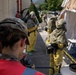 The image depicts what appears to be a HAZMAT or CBRN (Chemical, Biological, Radiological, Nuclear) training exercise or emergency response drill. Personnel in yellow hazmat suits with respirators and protective gear are operating near a decontamination tent setup. A military service member wearing a multicam cap with the name 'GONSALVES' is observing the operation in the foreground.