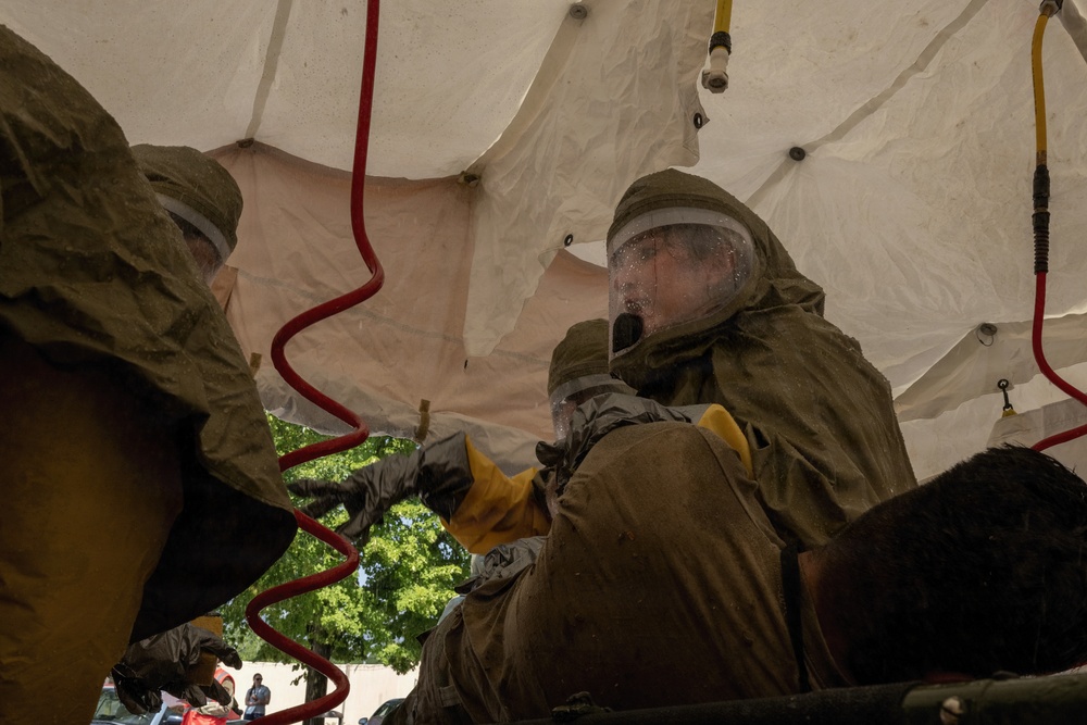 Two individuals wearing olive-drab CBRN (Chemical, Biological, Radiological, Nuclear) protective suits with full-face respirator visors and yellow rubber gloves are attending to a casualty under a white decontamination tent. The scene appears to be a hazmat or military emergency response exercise, with red corrugated hoses visible nearby suggesting decontamination equipment. A third person is partially visible in the background outside the tent.