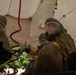 Two individuals wearing olive-drab CBRN (Chemical, Biological, Radiological, Nuclear) protective suits with full-face respirator visors and yellow rubber gloves are attending to a casualty under a white decontamination tent. The scene appears to be a hazmat or military emergency response exercise, with red corrugated hoses visible nearby suggesting decontamination equipment. A third person is partially visible in the background outside the tent.