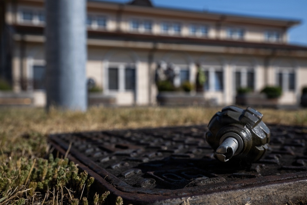 A close-up photograph shows what appears to be an inert or expended grenade body resting on a rusty metal drain grate near a grassy area. The foreground object is in sharp focus while a multi-story institutional or government-style building is visible in the soft-focus background. The scene appears to be an outdoor area, possibly a military base or public grounds.