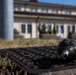 A close-up photograph shows what appears to be an inert or expended grenade body resting on a rusty metal drain grate near a grassy area. The foreground object is in sharp focus while a multi-story institutional or government-style building is visible in the soft-focus background. The scene appears to be an outdoor area, possibly a military base or public grounds.