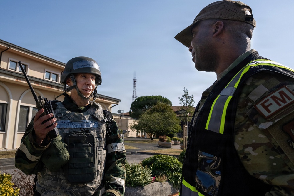 Two military personnel are engaged in a conversation outdoors on what appears to be a military installation. The individual on the left is wearing full tactical gear including a helmet and body armor and is holding a handheld radio. The individual on the right is wearing a high-visibility vest with a red 'FM' patch over a camouflage uniform and a tan cap, suggesting an exercise or evaluation role.