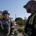 Two military personnel are engaged in a conversation outdoors on what appears to be a military installation. The individual on the left is wearing full tactical gear including a helmet and body armor and is holding a handheld radio. The individual on the right is wearing a high-visibility vest with a red 'FM' patch over a camouflage uniform and a tan cap, suggesting an exercise or evaluation role.