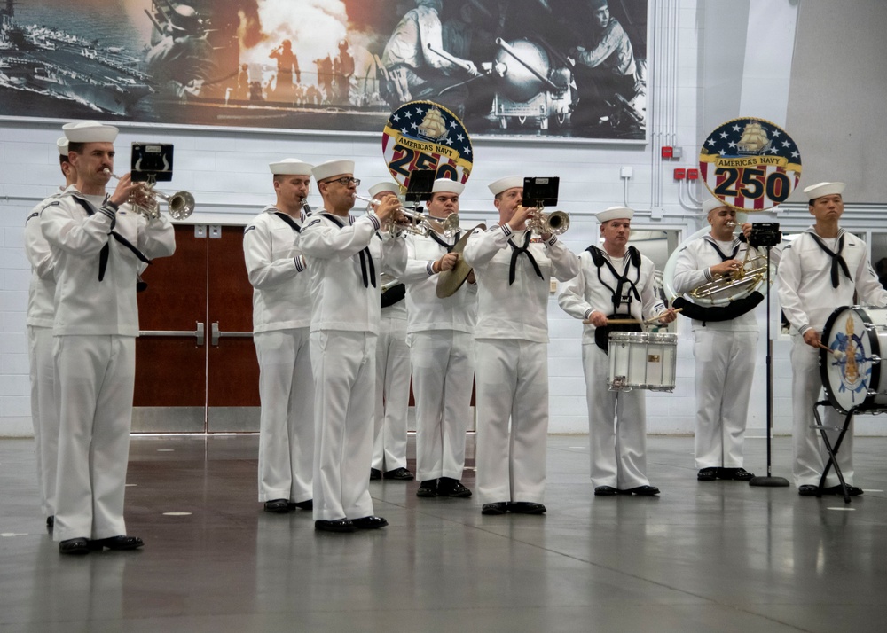 A U.S. Navy band performs indoors at what appears to be an official ceremony or event commemorating the Navy's 250th anniversary. The musicians are dressed in traditional white Navy dress uniforms and are playing brass instruments and percussion. The venue features large historical photographic murals on the walls and prominent 'America's Navy 250' commemorative emblems.