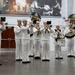A U.S. Navy band performs indoors at what appears to be an official ceremony or event commemorating the Navy's 250th anniversary. The musicians are dressed in traditional white Navy dress uniforms and are playing brass instruments and percussion. The venue features large historical photographic murals on the walls and prominent 'America's Navy 250' commemorative emblems.