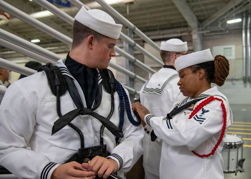 The image shows U.S. Navy personnel in dress white uniforms, with one sailor appearing to assist or inspect another sailor's uniform or equipment. The setting appears to be an indoor military facility, likely a hangar bay or drill hall. A drum is visible in the background, suggesting this may be a band or ceremonial unit preparation.