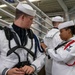 The image shows U.S. Navy personnel in dress white uniforms, with one sailor appearing to assist or inspect another sailor's uniform or equipment. The setting appears to be an indoor military facility, likely a hangar bay or drill hall. A drum is visible in the background, suggesting this may be a band or ceremonial unit preparation.