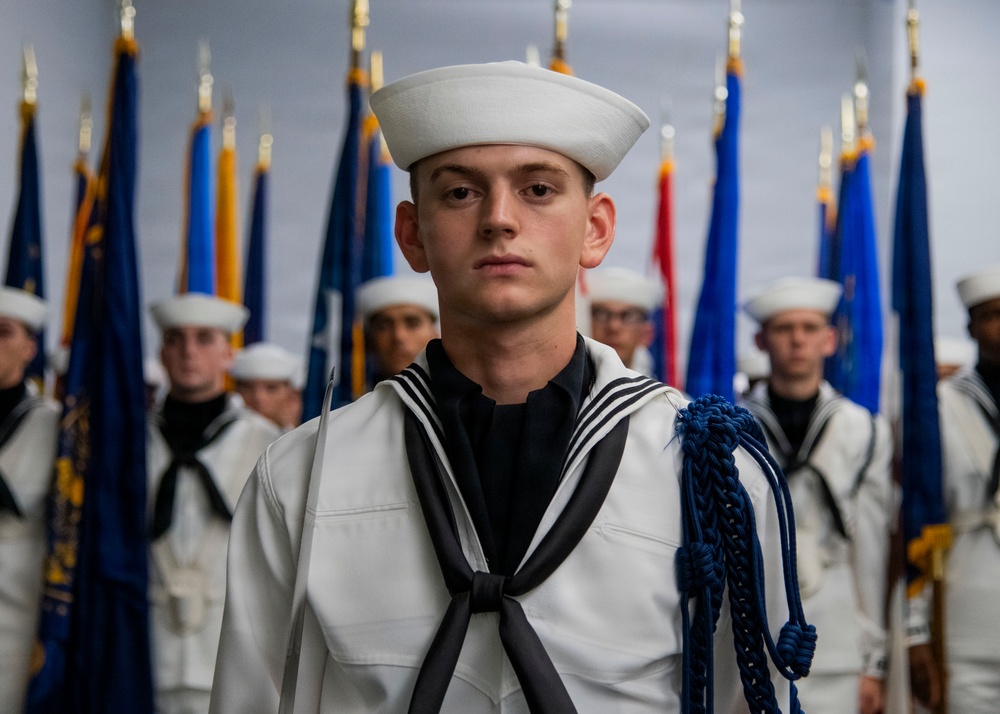 A young U.S. Navy sailor in dress white uniform stands at attention in the foreground, wearing a traditional Dixie cup white hat and a blue aiguillette on his right shoulder. Behind him, multiple sailors in identical dress whites are arranged in formation in front of a row of military unit flags with gold finials. The setting appears to be a formal military ceremony or graduation event.