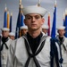 A young U.S. Navy sailor in dress white uniform stands at attention in the foreground, wearing a traditional Dixie cup white hat and a blue aiguillette on his right shoulder. Behind him, multiple sailors in identical dress whites are arranged in formation in front of a row of military unit flags with gold finials. The setting appears to be a formal military ceremony or graduation event.