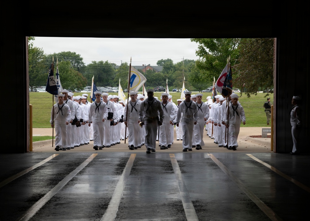 A large formation of U.S. Navy sailors in white dress uniforms is marching through a large open bay entrance, likely entering a drill hall or hangar. The formation is led by a drill instructor in gray physical training attire, with flag bearers carrying guidons on both flanks. The wet floor and overcast sky suggest recent rainfall during what appears to be a formal military ceremony or graduation event.