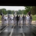 A large formation of U.S. Navy sailors in white dress uniforms is marching through a large open bay entrance, likely entering a drill hall or hangar. The formation is led by a drill instructor in gray physical training attire, with flag bearers carrying guidons on both flanks. The wet floor and overcast sky suggest recent rainfall during what appears to be a formal military ceremony or graduation event.