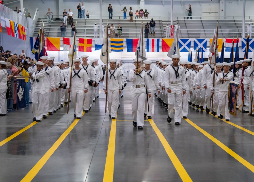 A large formation of U.S. Navy recruits in white dress uniforms marches in precise columns during what appears to be a boot camp graduation ceremony held indoors. Multiple color guard members carry flags and guidons at the front of the formation. Spectators and family members line the sides and upper balcony of the large drill hall or gymnasium.