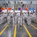 A large formation of U.S. Navy recruits in white dress uniforms marches in precise columns during what appears to be a boot camp graduation ceremony held indoors. Multiple color guard members carry flags and guidons at the front of the formation. Spectators and family members line the sides and upper balcony of the large drill hall or gymnasium.