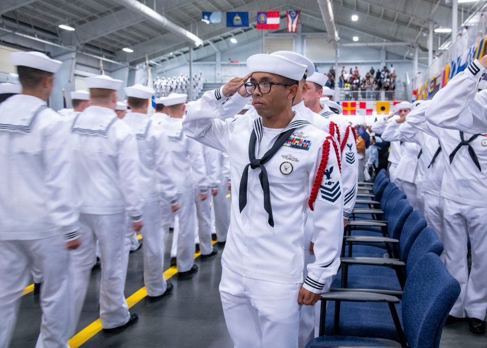 A formal U.S. Navy military ceremony taking place in a large indoor facility, likely a graduation or pass-in-review event. A prominent sailor in the foreground is rendering a salute while wearing dress white uniforms with ribbons, medals, and a red aiguillette, indicating a senior enlisted rank. Rows of sailors in matching dress whites are lined up in formation in the background.