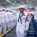 A formal U.S. Navy military ceremony taking place in a large indoor facility, likely a graduation or pass-in-review event. A prominent sailor in the foreground is rendering a salute while wearing dress white uniforms with ribbons, medals, and a red aiguillette, indicating a senior enlisted rank. Rows of sailors in matching dress whites are lined up in formation in the background.