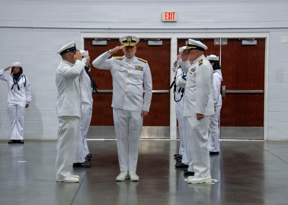A formal U.S. Navy military ceremony is taking place indoors, likely a change of command or official review. A senior officer in the center, distinguished by gold shoulder boards and a medal-laden uniform, is receiving salutes from personnel on both sides. The setting appears to be a gymnasium or large indoor hall with standard institutional features.