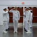 A formal U.S. Navy military ceremony is taking place indoors, likely a change of command or official review. A senior officer in the center, distinguished by gold shoulder boards and a medal-laden uniform, is receiving salutes from personnel on both sides. The setting appears to be a gymnasium or large indoor hall with standard institutional features.
