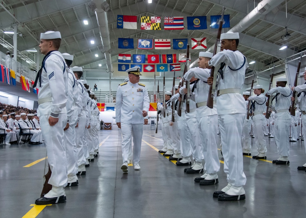A formal U.S. Navy graduation or pass-in-review ceremony taking place inside a large indoor drill hall or gymnasium. A senior officer, distinguished by gold shoulder boards and medal ribbons on a white dress uniform, walks between two rows of enlisted sailors in white dress uniforms holding rifles at attention. Numerous U.S. state flags are displayed hanging from the ceiling rafters above the formation.