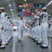 A formal U.S. Navy graduation or pass-in-review ceremony taking place inside a large indoor drill hall or gymnasium. A senior officer, distinguished by gold shoulder boards and medal ribbons on a white dress uniform, walks between two rows of enlisted sailors in white dress uniforms holding rifles at attention. Numerous U.S. state flags are displayed hanging from the ceiling rafters above the formation.