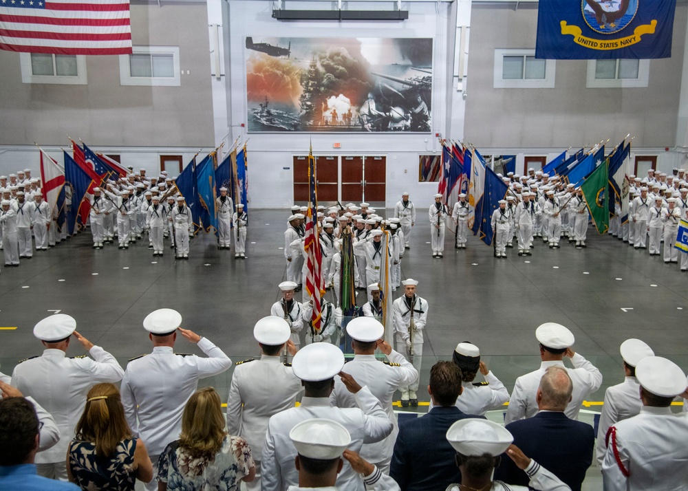 A formal United States Navy military ceremony is taking place inside a large indoor drill hall or gymnasium. Sailors in dress white uniforms are arranged in formation on both sides of the hall, while a color guard presents multiple state and organizational flags at the center. Officers and civilians in the foreground are rendering salutes toward the colors.