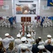 A formal United States Navy military ceremony is taking place inside a large indoor drill hall or gymnasium. Sailors in dress white uniforms are arranged in formation on both sides of the hall, while a color guard presents multiple state and organizational flags at the center. Officers and civilians in the foreground are rendering salutes toward the colors.