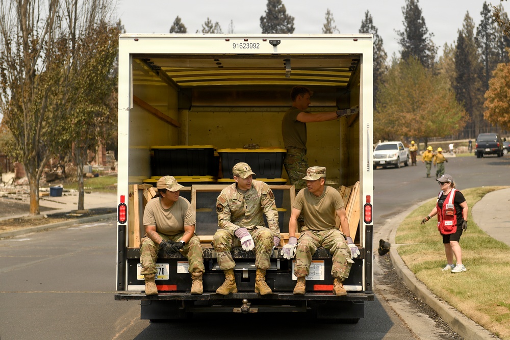 Military personnel in camouflage uniforms are seated at the rear of a box truck in what appears to be a wildfire disaster zone. The surrounding area shows significant fire damage with scorched trees and debris. Multiple response teams including military, firefighters in yellow gear, and a Red Cross volunteer are visible working in the area.