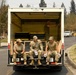 Military personnel in camouflage uniforms are seated at the rear of a box truck in what appears to be a wildfire disaster zone. The surrounding area shows significant fire damage with scorched trees and debris. Multiple response teams including military, firefighters in yellow gear, and a Red Cross volunteer are visible working in the area.