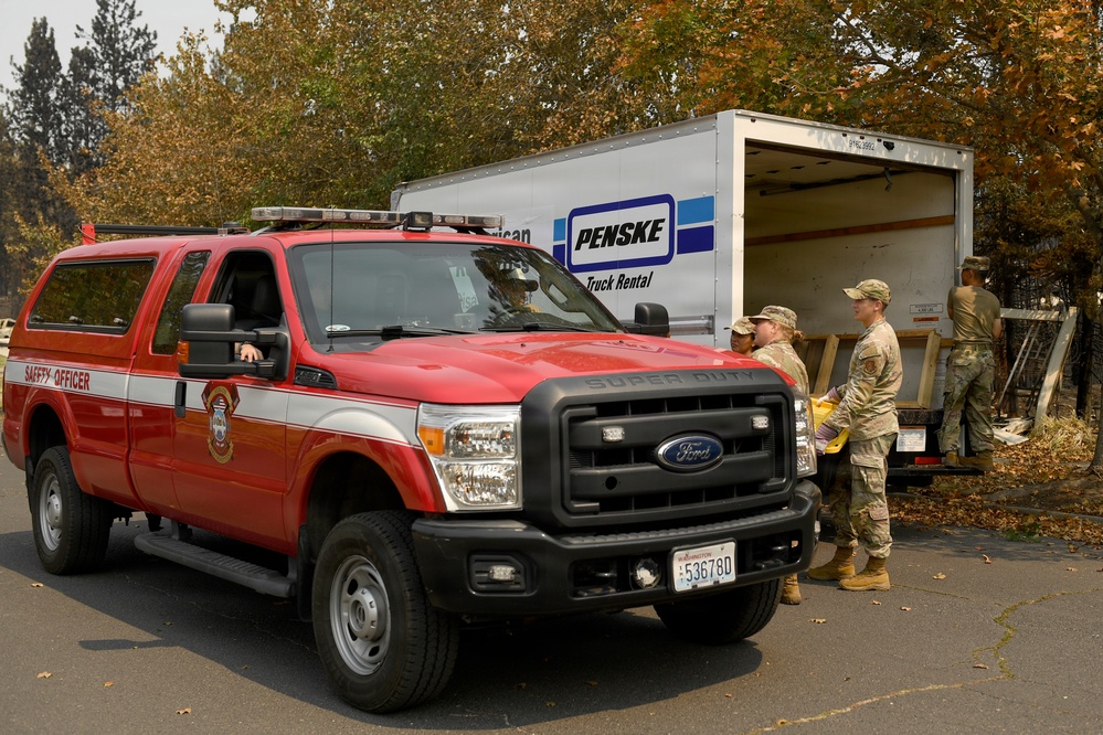 A red Ford Super Duty truck marked 'Safety Officer' is parked in front of an open Penske rental truck. Several individuals in military camouflage uniforms are gathered near the rear of the Penske truck, appearing to be loading or unloading materials. The scene is set in a wooded area with trees showing signs of fire damage or autumn foliage.