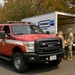 A red Ford Super Duty truck marked 'Safety Officer' is parked in front of an open Penske rental truck. Several individuals in military camouflage uniforms are gathered near the rear of the Penske truck, appearing to be loading or unloading materials. The scene is set in a wooded area with trees showing signs of fire damage or autumn foliage.