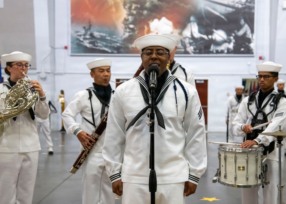 A U.S. Navy musical ensemble is performing indoors, likely at a military ceremony or event. A sailor in traditional white dress uniform stands at a microphone as a vocalist, while band members behind him play instruments including a French horn, clarinet, and snare drum. A large naval-themed mural is visible on the wall in the background.
