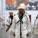 A U.S. Navy musical ensemble is performing indoors, likely at a military ceremony or event. A sailor in traditional white dress uniform stands at a microphone as a vocalist, while band members behind him play instruments including a French horn, clarinet, and snare drum. A large naval-themed mural is visible on the wall in the background.