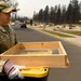 A U.S. Air Force service member named Brown is carrying a wooden-framed sifting screen and a yellow container in what appears to be a fire-devastated neighborhood. The surrounding area shows extensive destruction with burned debris, ash piles, and charred trees consistent with a major wildfire aftermath. The soldier appears to be involved in search and recovery operations.